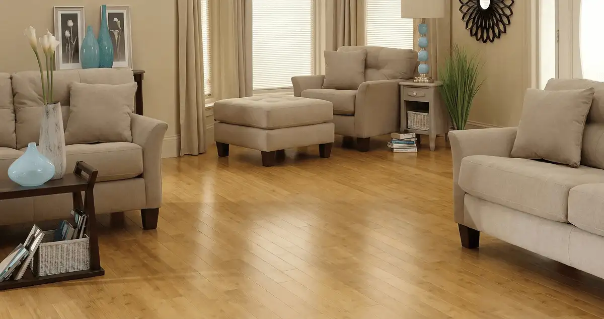 A living room with professionally installed bamboo flooring, beige sofas, a matching ottoman, a side table with a blue lamp, vases, framed pictures, and large windows with blinds and curtains in Scottsdale, AZ